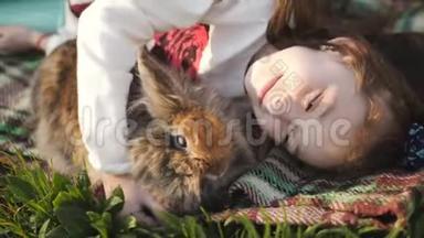 小女孩和棕色兔子躺在绿色的草坪上拥抱。 女孩玩最喜欢的宠物。 一只兔子。