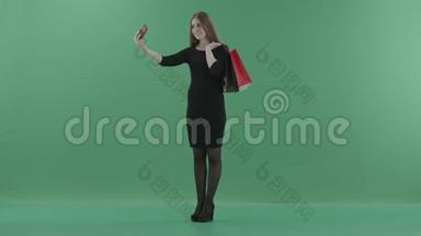穿着黑色<strong>小裙子</strong>的漂亮女人拿着购物袋在手机上自拍。 她是
