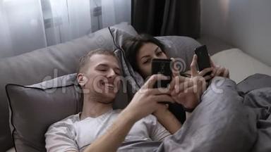 年轻的已婚夫妇在床上躺着，互相微笑时使用手机。 快乐的生活方式，情侣，床