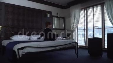 在一家舒适的小旅馆里，一个年轻疲惫的女人坐在一张漂亮的宽床上，对面是<strong>落地窗</strong>和百叶窗。