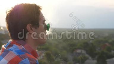 戴墨镜的英<strong>俊男</strong>子站在屋顶，在城市景观背景下吹烟。 年轻人休息