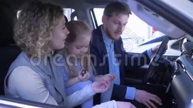 家庭在汽车销售中心，快乐的微笑爸爸妈妈和小女孩回顾汽车和挥动钥匙坐着