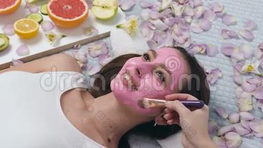 可爱的白种人女人在健康中心放松的肖像，而美容师使用化妆刷敷粉红色面具。