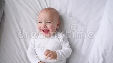 一个穿着白色t恤的孩子躺在一张白色的床上，看着摄像机，慢镜头地笑着，俯视着