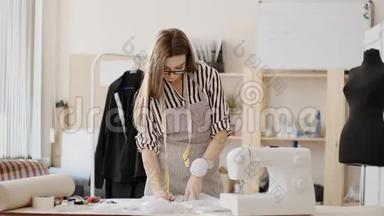 女裁缝、眼镜和围裙，站在缝纫机附近的办公桌后面，在缝纫机上做铅笔标记