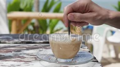 在大海和绿色植物的背景下喝杯咖啡。 特写男手搅动咖啡.. 在海滩上吃早餐
