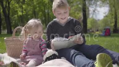 <strong>年轻</strong>的金发男孩坐在公园的毯子上，给她穿着粉红色裙子的妹妹读这本书，坐在附近。 这<strong>就是</strong>