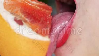 女人`舌头舔葡萄柚。 一个女孩的肖像，明亮的红色嘴唇舔一个粉红色的葡萄柚。 关门