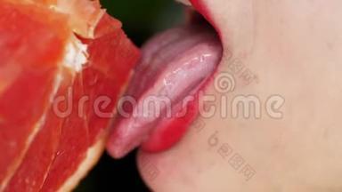 女人`舌头舔葡萄柚。 一个女孩的肖像，明亮的红色嘴唇舔一个粉红色的葡萄柚。 关门