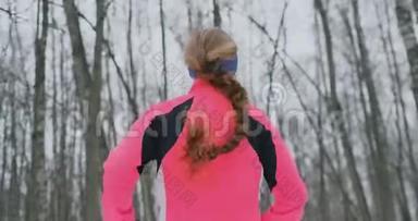 冬天，一个年轻的女人穿着一件粉红色的夹克穿过公园。 从后面看