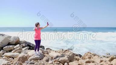 年轻漂亮的适合女孩拿着电话和自拍照片，而站在岩石海岸。 海浪拍打着岩石