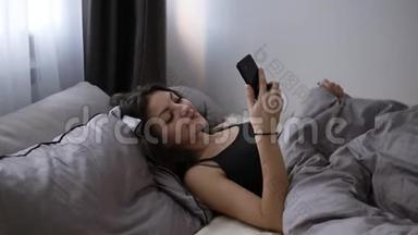 年轻开朗的黑发女孩<strong>躺</strong>在毯子下，在<strong>床上看</strong>着她的<strong>手机</strong>。 白天放松时间，