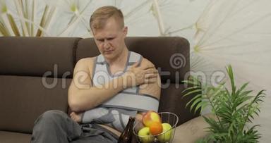 一个<strong>手</strong>臂<strong>骨折</strong>的痛苦男人，戴着臂架坐在沙发上喝啤酒