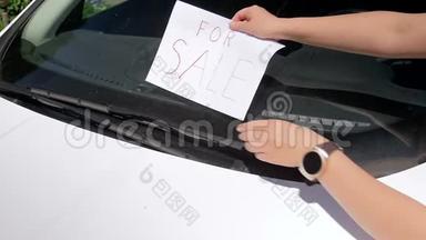 特写慢动作视频，年轻女子在她的汽车屏幕雨刷下放置待售标志。 女人出卖她