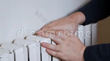 一个戴帽子的人把手放在靠近墙壁的暖气片上取暖。 公寓里冷，<strong>供暖</strong>系统差