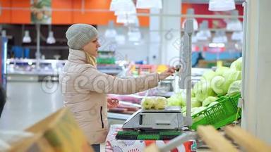 穿着冬装的女人在超市秤着苹果。 她选择水果的代码和价值。