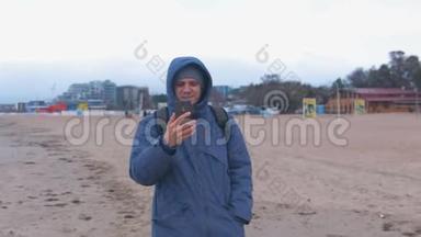 男人博主穿着蓝色羽绒服在海边沙滩上散步，在手机上聊天。