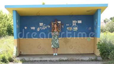 站在彩绘<strong>公交</strong>车站的一个穿着连衣裙的非常时尚漂亮的女孩。她在路边吃纸杯里的种子。