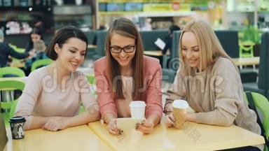 快乐的年轻女士们一起使用智能手机，看着屏幕，微笑着，笑着坐在咖啡馆购物