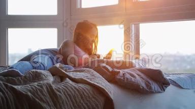 躺在床上的年轻美女在令人惊叹的日落中使用手机，并具有镜头耀斑效果。 慢慢慢慢
