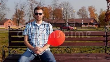 戴着黑色眼镜的野蛮年轻人坐在一张带着红色气球的长凳上，看着相机，复制空间，慢动作