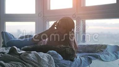 年轻女子戴着耳机，与她可爱的缅因州浣熊猫在床上放松，听着窗外的音乐唱歌