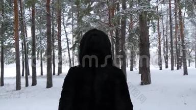 穿着黑色毛皮大衣的女人穿过冬天的森林或公园。 镜头移向在雪地里行走的女孩身后..