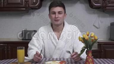 漂亮的年轻人穿着<strong>白色浴袍</strong>坐在厨房的桌子旁，享受着早餐的香气。 煎锅