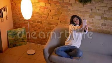 漂亮的黑发女商人坐在沙发上，在家里愉快地在智能手机上制作自拍照片。