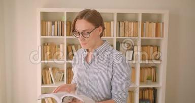 一位年轻貌美的白种<strong>人</strong>男学生戴着眼镜看着<strong>图书馆</strong>里的摄像机看着一本书