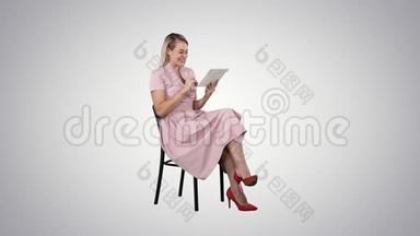 有趣的漂亮年轻女人穿着粉红色的连衣裙坐在椅子上，在渐变背景上使用平板电脑。