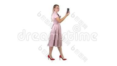 穿着粉色衣服的年轻女子在白色背景下行走时使用智能手机录制视频博客。