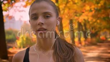 一个年轻的白种人女孩的特写肖像，她带着耳机观看镜头，试图在跑步后喘口气。