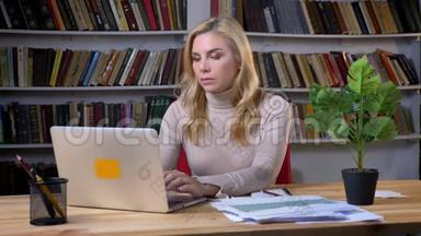 金发白种人女人在笔记本电脑<strong>前</strong>工作，在<strong>书架</strong>背景上做笔记。