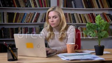 白种人金发中年妇女在<strong>书架</strong>背景上的笔记本电脑<strong>前</strong>打哈欠的肖像。
