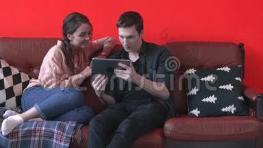 一对幸福的年轻夫妇坐在家里的棕色沙发上，看着平板电脑屏幕。 库存数据