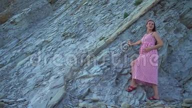 年轻的怀孕女孩穿着条纹红白连衣裙。 一个女孩站在岩石山脚下，<strong>抚摸</strong>着她的肚子。
