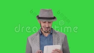 英俊的微笑阿拉伯男人戴着帽子走路，用平板电脑在绿色屏幕上，Chroma键。