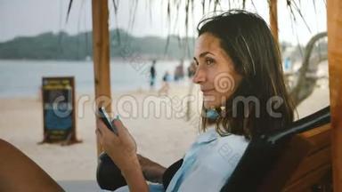 侧面拍摄的年轻快乐微笑的旅游妇女与饮料和智能手机休息，环顾海滩椅子。