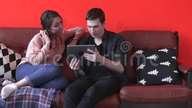 一对幸福的年轻夫妇坐在家里的棕色沙发上，看着平板电脑屏幕。 库存数据