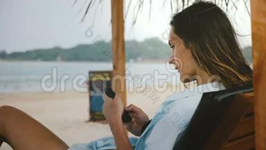 侧面拍摄年<strong>轻放</strong>松的自由职业者妇女使用智能手机购物应用程序在休闲海滩椅子与海景。