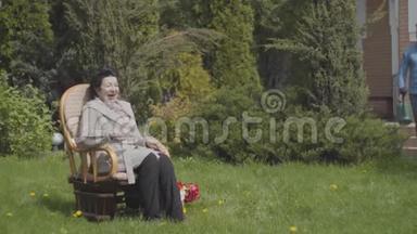 肖像成熟的女士坐在摇椅上享受阳光。 成年孙子带着<strong>水缸</strong>来
