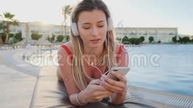 年轻迷人的女士穿着红色比基尼和白色耳机，听着智能手机里的音乐，一边躺着一边唱歌