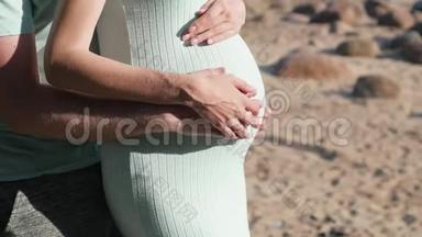 年轻的孕妇和丈夫走在海边。 一家人期待着一个婴儿沿着海岸散步。 腹部特写