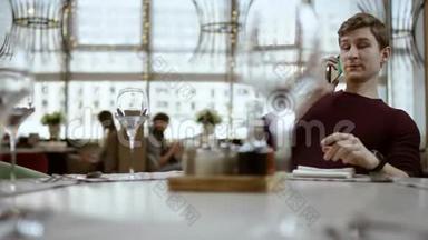 年轻英俊的自由职业者带着笔记本电脑在餐桌上工作，在电话上输入一条信息。 自由职业者的工作概念