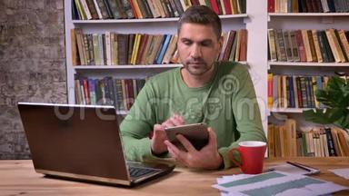 商人坐在笔记本电脑前，喝着咖啡，在平板电脑上滚动页面。 书架上的书架