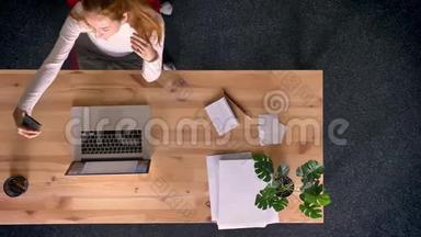 多莉拍摄，从上往下看，红头发的女人在办公室里用智能手机制作自拍照片。