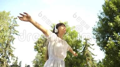 一个戴着耳机的快乐女孩微笑着，带着<strong>手机</strong>在公园里跳舞和<strong>旋转</strong>。 美丽的女孩听