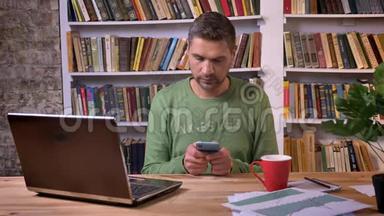 商人坐在笔记本电脑前，在手机上浏览消息。 背景书架