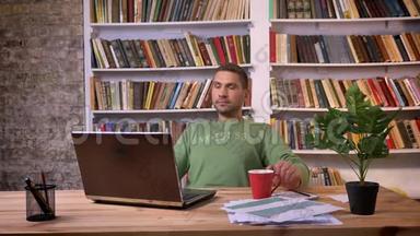 商人在笔记本电脑上工作，喝咖啡，并试图集中精力在<strong>办公室</strong>。 <strong>背景书架</strong>